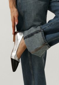 Zilverkleurige metallic platte schoen met een puntige zwarte neus, gedragen met versleten blauwe jeans met een brede omslag en een rafelige zoom.
