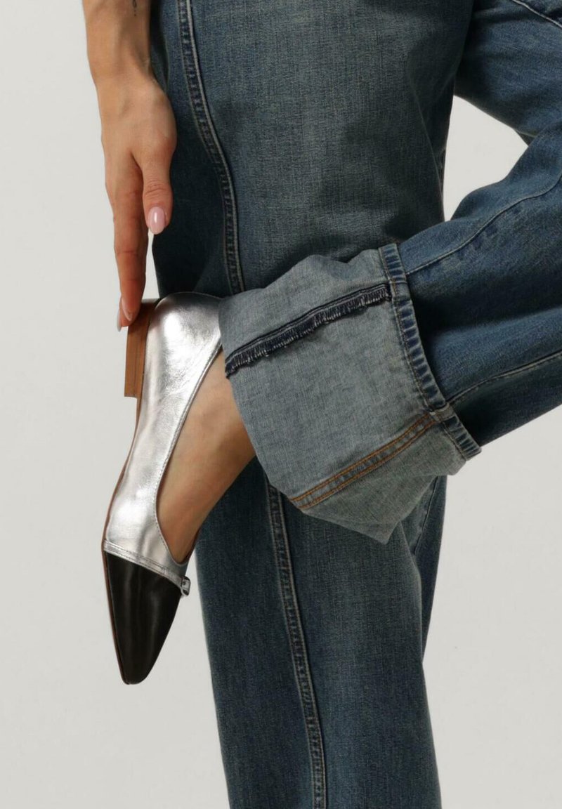 Zilverkleurige metallic platte schoen met een puntige zwarte neus, gedragen met versleten blauwe jeans met een brede omslag en een rafelige zoom.