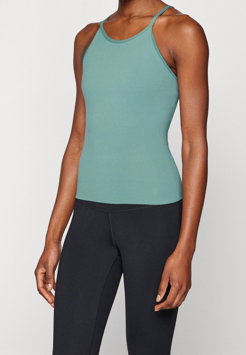 Una camiseta sin mangas ajustada de tela teal con una textura suave y tirantes finos, combinada con leggings negros que tienen costuras planas.