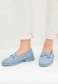 Lichtblauwe loafers met geperforeerde textuur, platte rubberen zool en decoratieve kettingdetails op de bovenkant. Opgerolde witte broek laat het schoeisel zien.