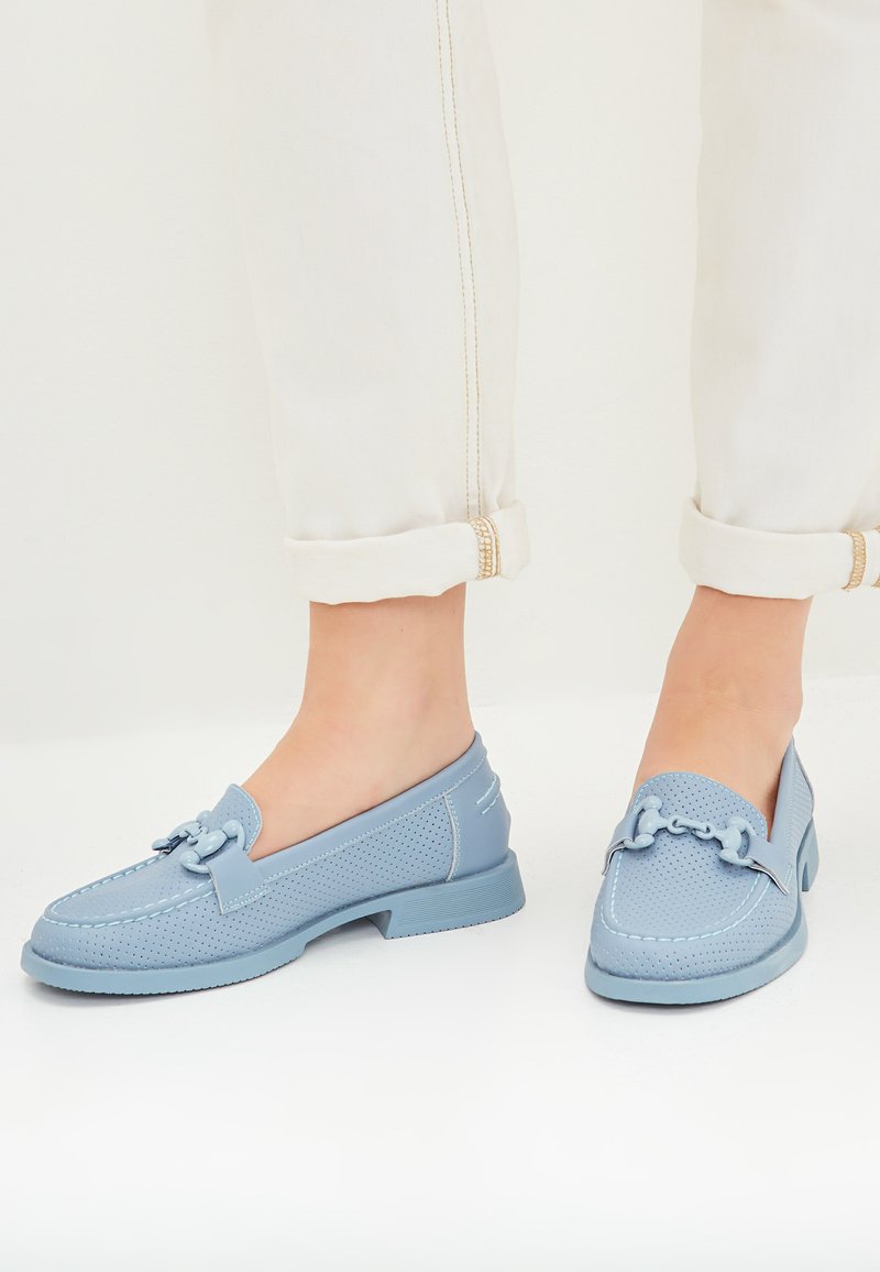 Lichtblauwe loafers met geperforeerde textuur, platte rubberen zool en decoratieve kettingdetails op de bovenkant. Opgerolde witte broek laat het schoeisel zien.
