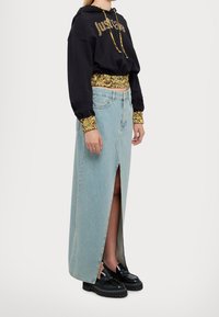 Persona che indossa un hoodie cropped nero con stampa dorata e una lunga gonna di jeans blu chiaro con spacco frontale, abbinata a mocassini neri chunky.