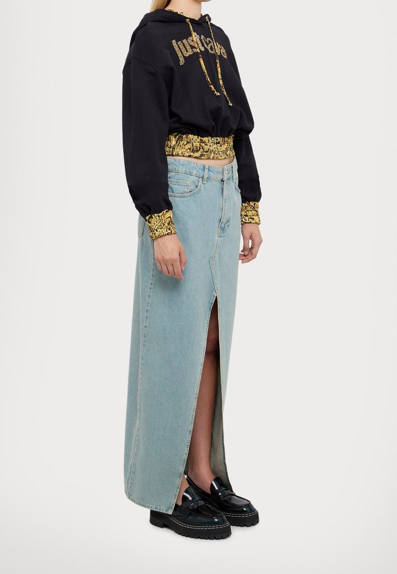 Persona che indossa un hoodie cropped nero con stampa dorata e una lunga gonna di jeans blu chiaro con spacco frontale, abbinata a mocassini neri chunky.