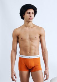 Πορτοκαλί βαμβακερά μποξεράκια με λευκή μέση που φέρει το λογότυπο "TOMMY HILFIGER", παρουσιάζονται σε αντρικό μοντέλο πάνω σε απλό φόντο.