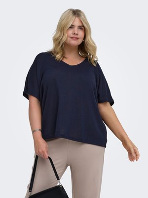 Blonde vrouw draagt een losse donkerblauwe top met korte mouwen en beige broek, houdt een zwarte handtas naast zich tegen een witte achtergrond.