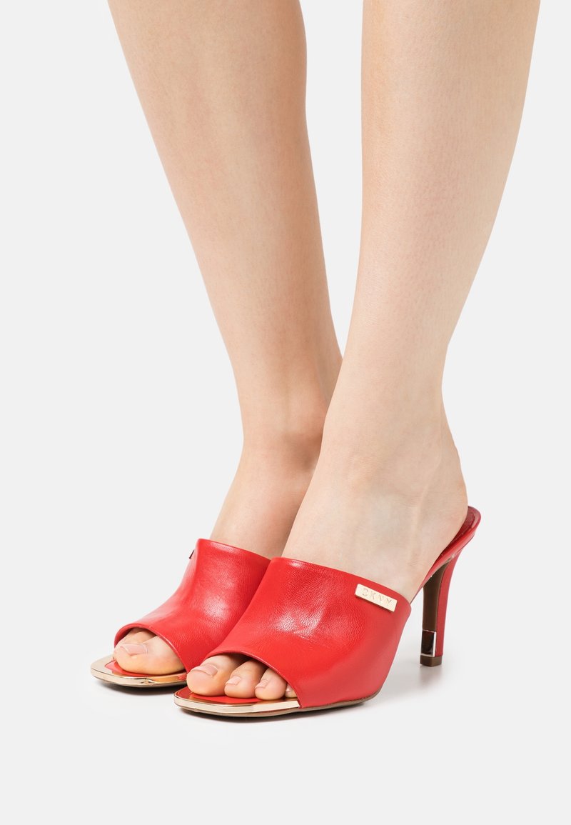 Mules ouvertes en cuir rouge avec une texture lisse, présentant un accent doré et un talon fin. Le design comprend une large lanière supérieure.