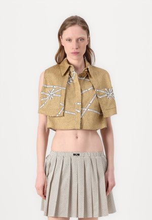 Golden cropped shirt with short sleeves featuring contrasting white ribbon patterns. Worn over a pleated, polka-dotted white skirt.