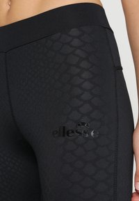 Czarne legginsy z teksturowanym wzorem w łuski, elastycznym pasem w talii oraz błyszczącym czarnym logo po lewej stronie.