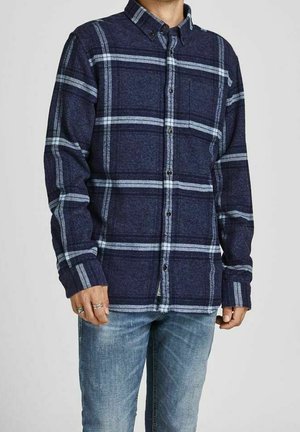 Homme portant une chemise à carreaux bleu marine et bleu clair à manches longues boutonnée, avec un jean clair, debout devant un fond uni.