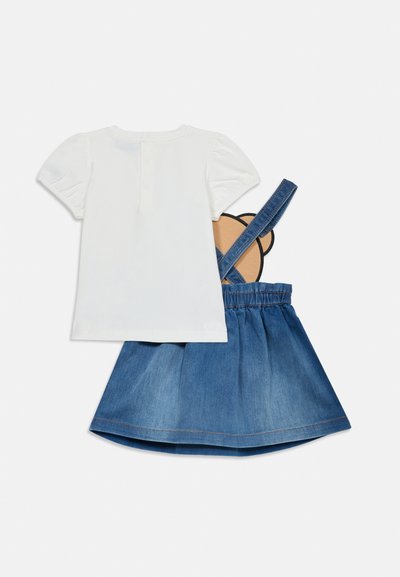 Chemise blanche à manches courtes associée à une jupe salopette en denim bleu avec un motif d'oreilles d'ours sur les bretelles arrière.