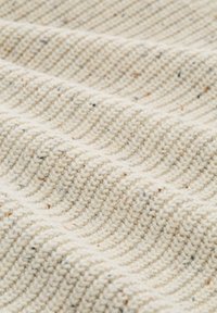 Gestructureerde crèmekleurige stof met een golvend patroon, voorzien van kleine vlekjes bruin. Het materiaal lijkt dik en geweven, waardoor een ribbelachtige uitstraling ontstaat.