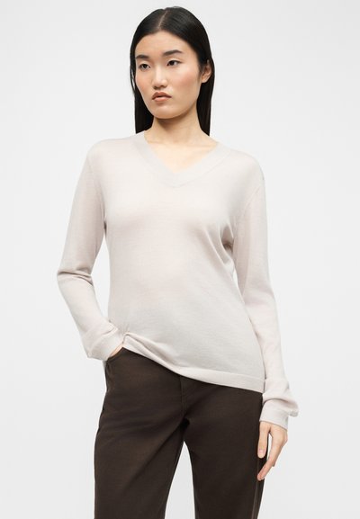 Γυναίκα με μακριά σκουρόχρωμα μαλλιά που φορεί ανοιχτό μπεζ πουλόβερ με V και σκούρο καφέ παντελόνι, στέκεται με το ένα χέρι στην τσέπη μπροστά σε λευκό φόντο.