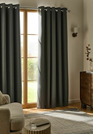 Closed dark gray curtains hang over a wooden framed glass door in a cozy living room with a beige armchair and round side table.
