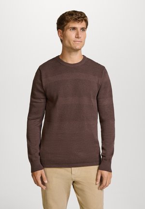 Uomo che indossa un maglione di lana marrone scuro a trama e pantaloni beige, in piedi davanti a uno sfondo chiaro uniforme, guardando leggermente verso destra.
