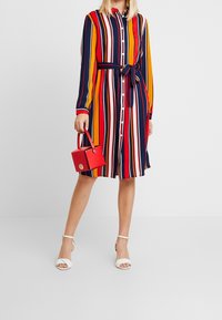 Robe à boutons rayée avec des lignes verticales rouges, orange, marine et blanches, dotée d'une ceinture nouée à la taille. Accompagnée d'un petit sac à main rouge.