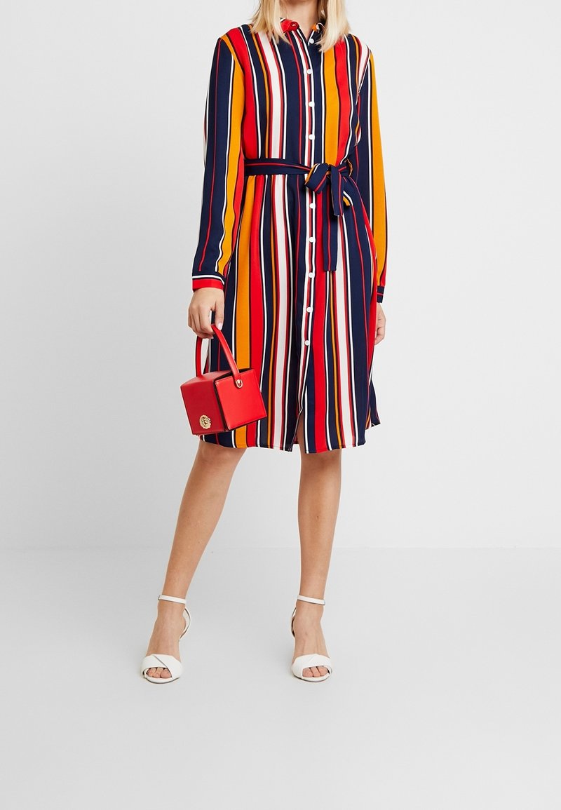 Robe à boutons rayée avec des lignes verticales rouges, orange, marine et blanches, dotée d'une ceinture nouée à la taille. Accompagnée d'un petit sac à main rouge.