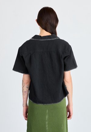 Persoon met lang donker haar in een staart draagt een zwart topje met korte mouwen met witte stiksel en een groene getextureerde rok, gezien van achteren.