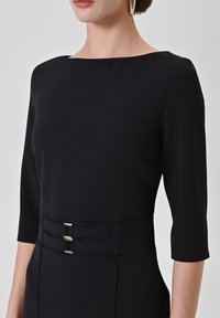 Robe noire à manches trois-quarts, dotée d'un col bateau et d'une taille cintrée soulignée par trois détails en métal.