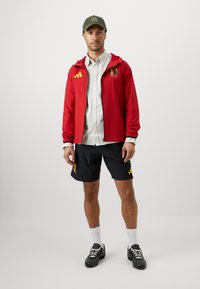 Κόκκινο ελαφρύ μπουφάν με κίτρινες λεπτομέρειες και βελγικό έμβλημα, συνδυασμένο με μαύρες σορτς που περιέχουν λογότυπα. Το σύνολο ολοκληρώνει ένα πράσινο καπέλο.