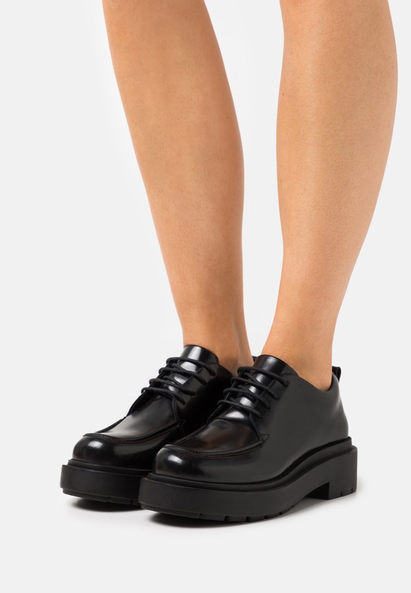 Chaussures noires en cuir brillant à lacets avec semelles épaisses portées sur des jambes nues sur un fond blanc.
