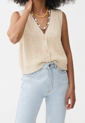 Personne portant un gilet beige sans manches en tricot boutonné, jean taille haute bleu clair, colliers de perles jaunes et coquillages, main près du visage.