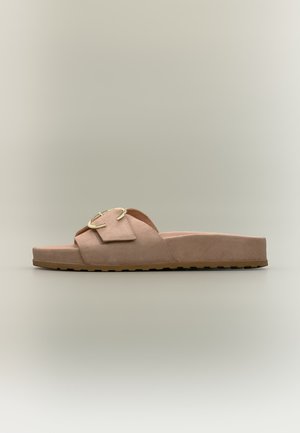 Sandale beige à enfiler avec une large bride et une grosse boucle dorée sur une semelle plate basse, sur fond neutre.