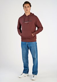 Felpa corta bordeaux con tasca frontale e cappuccio con cordino; presenta la scritta "COPENHAGEN" in testo nero. Abbinata a jeans blu e sneaker bianche.