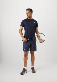 Άνδρας με ναυτικά αθλητικά μπλούζα και σορτς κρατάει μια ρακέτα τεννίς, όρθιος σε φόντο λευκού δαπέδου και τοίχου.