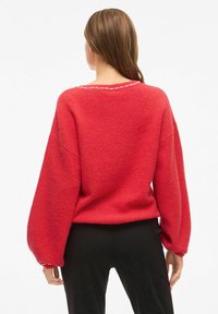 Maglione rosso, sovradimensionato, con una finitura testurizzata, maniche a palloncino e un colletto rotondo adornato da piccole perline lungo il bordo.
