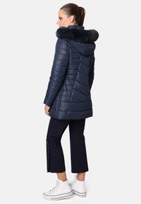 Abrigo acolchado azul marino con capucha de piel sintética, diseño acolchado y mangas largas. Combinado con pantalones negros cortos con detalles de botones y zapatillas blancas.