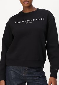 Sudadera negra hecha de una mezcla de algodón, con cuello redondo, puños y dobladillo acanalados, y logo en blanco "TOMMY HILFIGER EST. 1985".