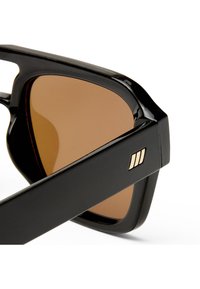 Μαύρα γυαλιά ηλίου με χοντρά ορθογώνια πλαίσια και φακούς με καφέ απόχρωση, με μικρή χρυσή λεπτομέρεια τριών ριγών στο μπράτσο του σκελετού.