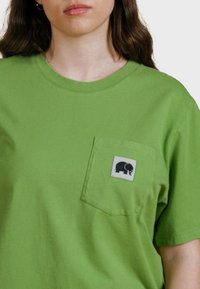 Camiseta de algodón verde con cuello redondo y mangas cortas. Presenta un bolsillo frontal con un parche gris que muestra un gráfico de un elefante negro.