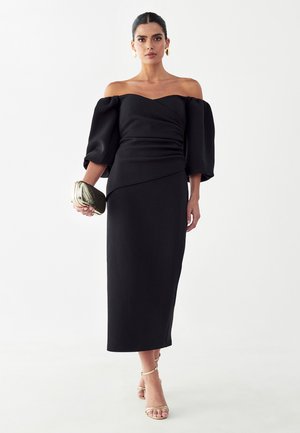 Žena v čiernych šatách s odhalenými ramenami a nafúknutými rukávmi, drží zelené prešívané psaníčko, nosí zlaté kruhové náušnice a sandále na tenkých remienkoch.