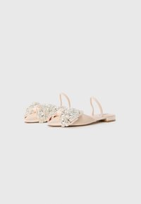 Light pink flat slingback shoes with pointed toes, adorned with large, sparkling crystal embellishments on the front bows.