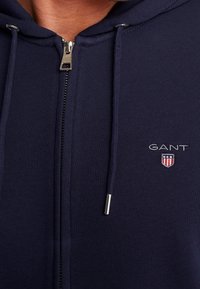 Navy modrý zipový svetr vyrobený z hladké tkaniny má stříbrný zip, šňůrky a vyšívané logo „GANT“ se štítem na hrudi.