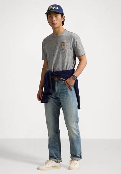 Szürke gyapjú t-shirt hímzett logóval, kék farmernadrág, tengerészkék pulóver a derekon, fehér cipő és kék sapka felirattal. Klasszikus laza öltözet.