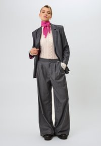 Harmaa raidallinen puku, jossa on leveät housut ja siihen sopiva bleiseri. Yläosana on pitsipaita vaaleanpunaisella neulotulla huivilla. Mustat jalkineet.