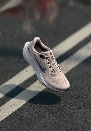 Lyserød Nike løbesko med sort Swoosh-logo, svæver over sort asfalt med hvide vejmarkeringer.