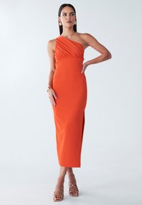 Ein schulterfreies oranges Kleid mit gerafftem Oberteil, enganliegender Silhouette und einem hohen Schlitz bis zum Oberschenkel. Getragen mit goldenen Riemchenschuhen und minimalem Schmuck.