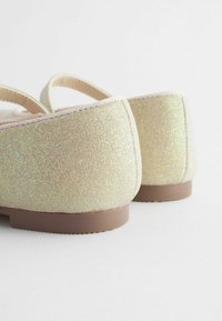 Pale gold színű, fényesen csillogó anyagból készült balerina cipők, rövid oldalakkal, kerek orral és lapos barna talppal.