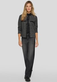 Graue, strukturierte Stoffjacke mit einem Reißverschluss vorne und zwei Taschen, kombiniert mit locker sitzenden dunklen Jeans und schwarzen Schuhen.