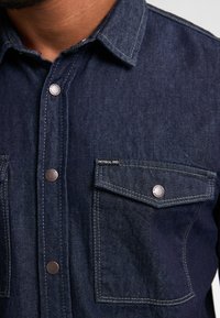 Camisa de mezclilla en azul oscuro con costuras en contraste, botones a presión y dos bolsillos en el pecho. Presenta la etiqueta de la marca en un bolsillo.