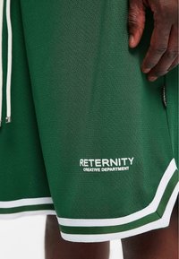 Zeleni mrežasti košarkaški šorc s bijelim špagama, s bijelim logotipom koji glasi "RETERNITY STVARALAŠKI ODJEL" blizu rubnog dijela i prugastim naglascima.