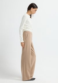 Frau mit dunkelhaarigen, die eine weiße Bluse mit Rüschenärmeln, beigen, hoch taillierten weiten Hosen und schwarzen spitzen Schuhen trägt, steht seitlich.