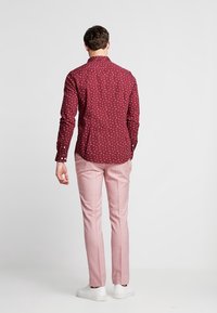 Camicia button-up bordeaux con motivi astratti bianchi, abbinata a pantaloni su misura rosa chiaro e scarpe bianche, visto da dietro.