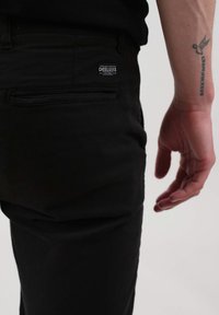 Pantalons en coton noirs avec une poche arrière visible et une étiquette. Main reposant à côté de la poche. Texture lisse, design ajusté.