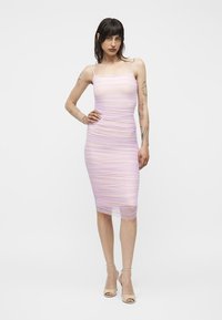 Femme aux cheveux foncés et tatouages portant une robe midi lavande cintrée froncée et des sandales à talons beiges, debout avec une main sur la hanche.
