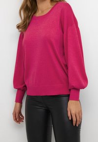 Pull en tricot fuchsia avec un col rond, des manches longues bouffantes et des poignets côtelés. Porté avec un pantalon noir ajusté.