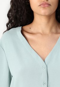 Blusa verde chiaro con scollo a V e tre bottoni. Il tessuto appare morbido con una leggera texture. Vista ravvicinata della parte superiore del torso.